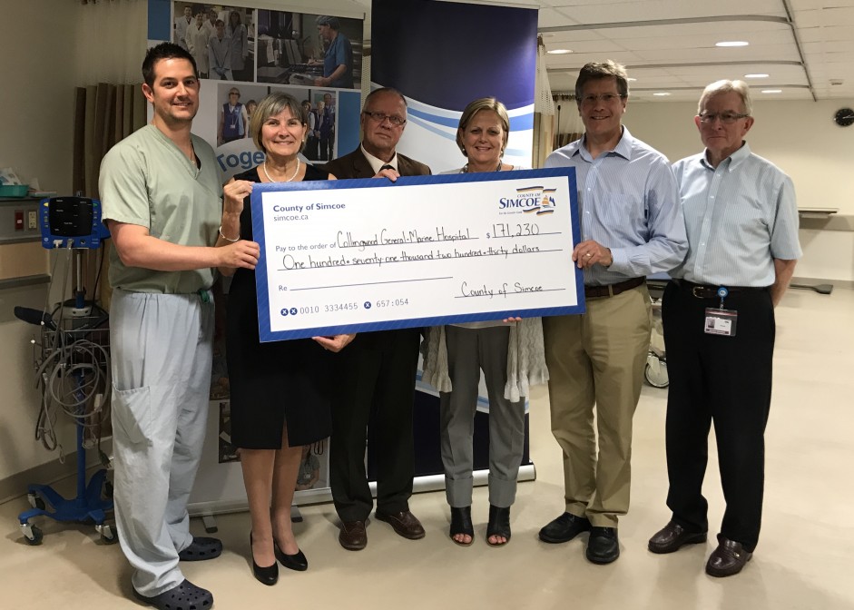 A group of men and women stand side by side inside a hospital recovery room holding an oversized donation cheque.
